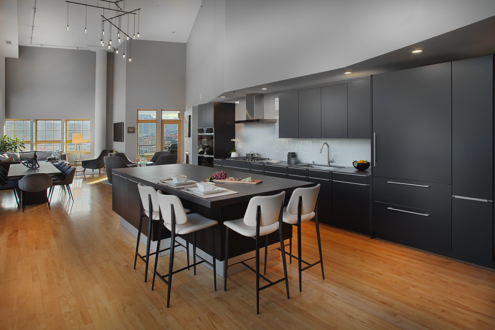 Photo of a large contemporary l-shaped open plan kitchen in Minneapolis with an undermount sink, flat-panel cabinets, black cabinets, quartz benchtops, white splashback, glass sheet splashback, panelled appliances, light hardwood floors, with island and black benchtop.