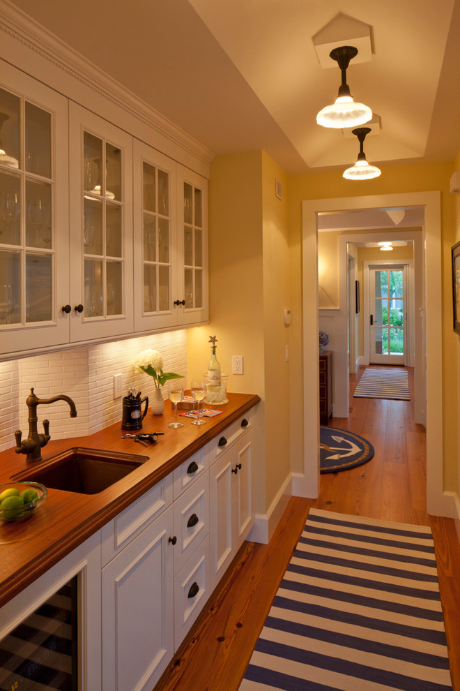Mid-sized beach style single-wall medium tone wood floor and brown floor wet bar photo in Boston with an undermount sink, glass-front cabinets, white cabinets, wood countertops, white backsplash and brown countertops