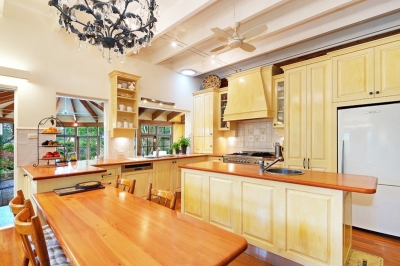 This is an example of a mid-sized country l-shaped open plan kitchen in Sydney with a single-bowl sink, raised-panel cabinets, yellow cabinets, wood benchtops, beige splashback, stone tile splashback, medium hardwood floors and multiple islands.