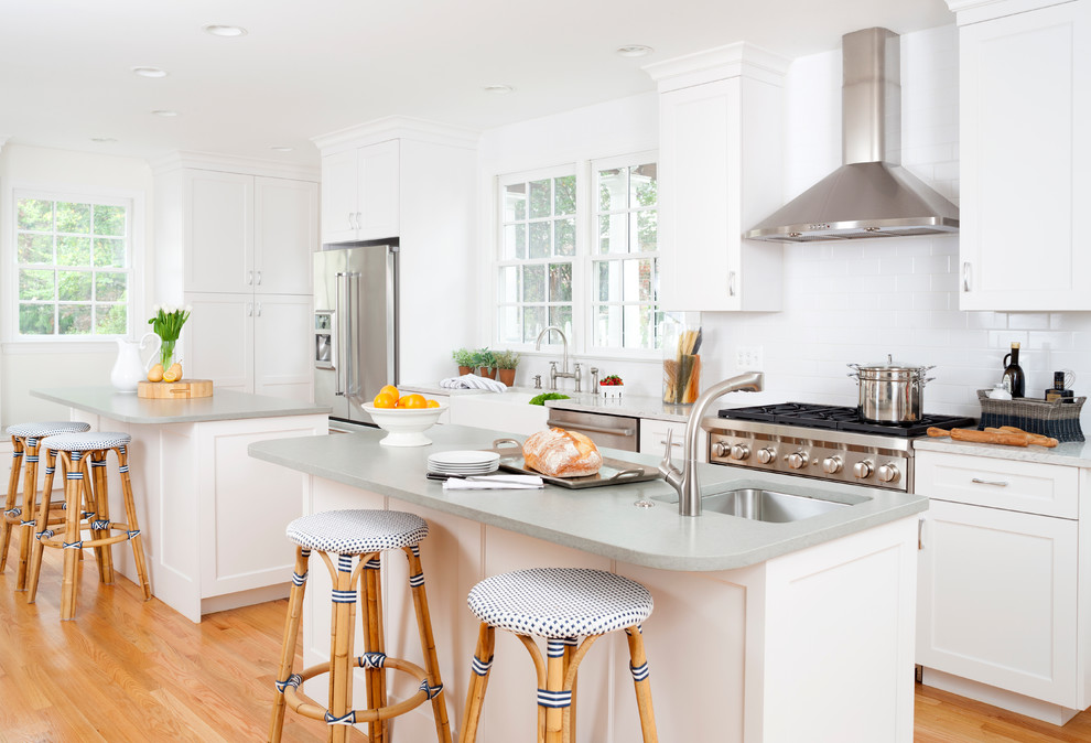 This is an example of a transitional galley kitchen in Baltimore with a farmhouse sink, shaker cabinets, white cabinets, white splashback, subway tile splashback, stainless steel appliances, light hardwood floors and multiple islands.