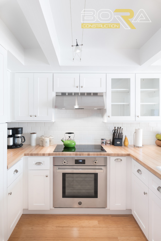 Example of a small transitional u-shaped medium tone wood floor and beige floor eat-in kitchen design with a double-bowl sink, shaker cabinets, white cabinets, wood countertops, white backsplash, ceramic backsplash, stainless steel appliances and a peninsula