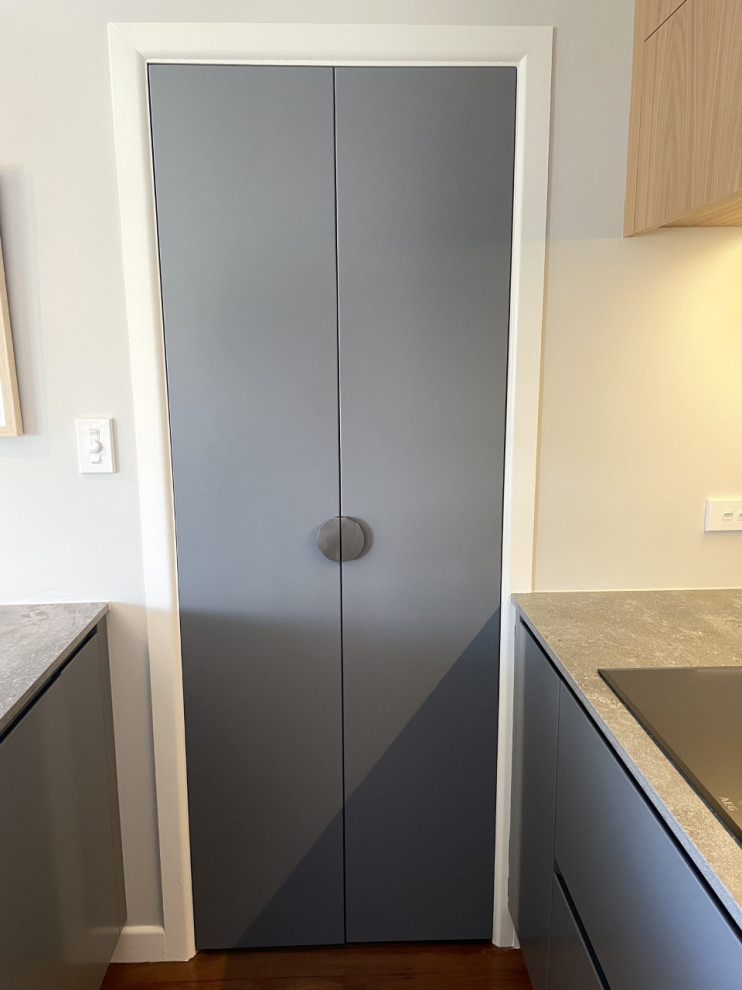 This is an example of a small contemporary galley kitchen/diner in Auckland with a double-bowl sink, flat-panel cabinets, grey cabinets, grey splashback, porcelain splashback, black appliances, light hardwood flooring, a breakfast bar, brown floors and grey worktops.