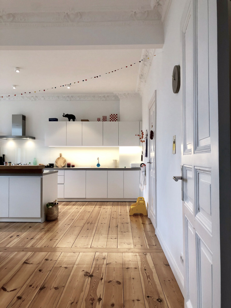 Photo of a large contemporary single-wall eat-in kitchen in Berlin with a drop-in sink, flat-panel cabinets, white cabinets, limestone benchtops, white splashback, glass sheet splashback, black appliances, light hardwood floors, with island, brown floor and grey benchtop.