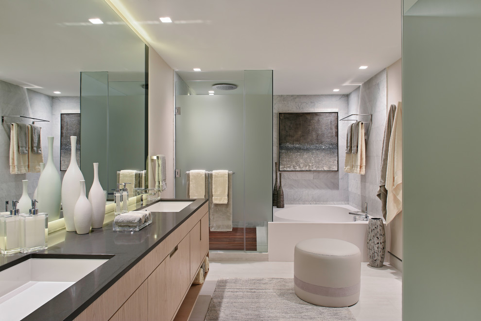 This is an example of a contemporary bathroom in Miami with flat-panel cabinets, light wood cabinets, a corner tub, a corner shower, gray tile, beige walls, an undermount sink, beige floor, a hinged shower door and black benchtops.