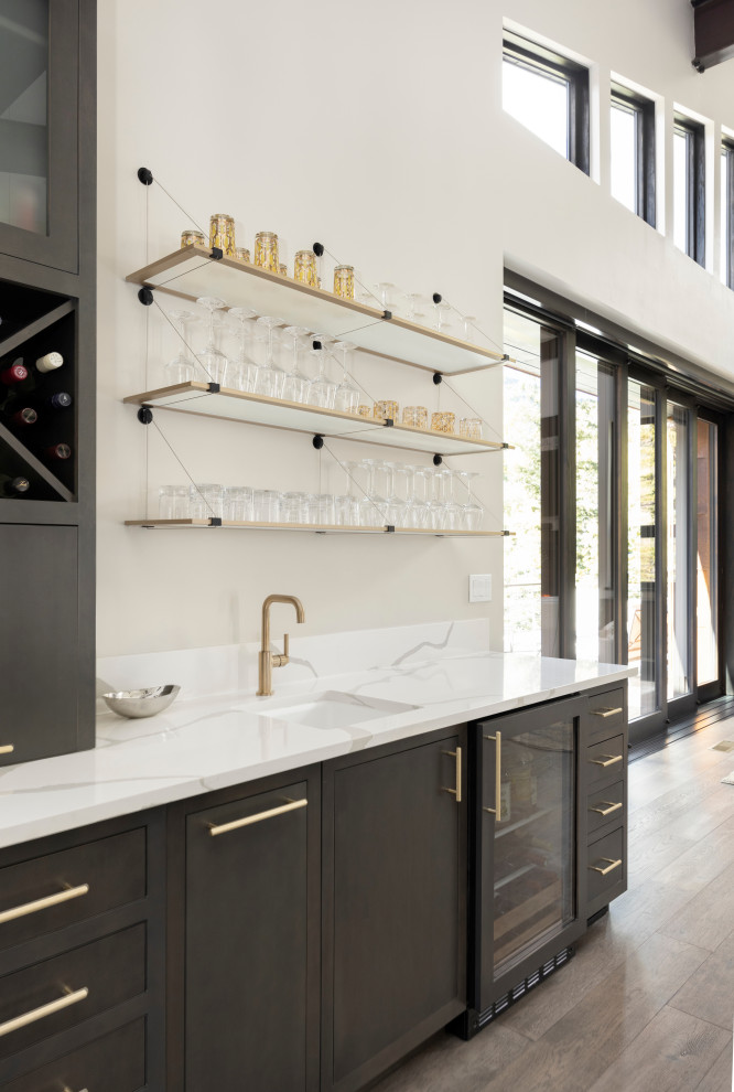 Photo of a large contemporary u-shaped wet bar in Denver with a drop-in sink, shaker cabinets, grey cabinets, quartzite benchtops, white splashback, engineered quartz splashback, medium hardwood floors, brown floor and white benchtop.