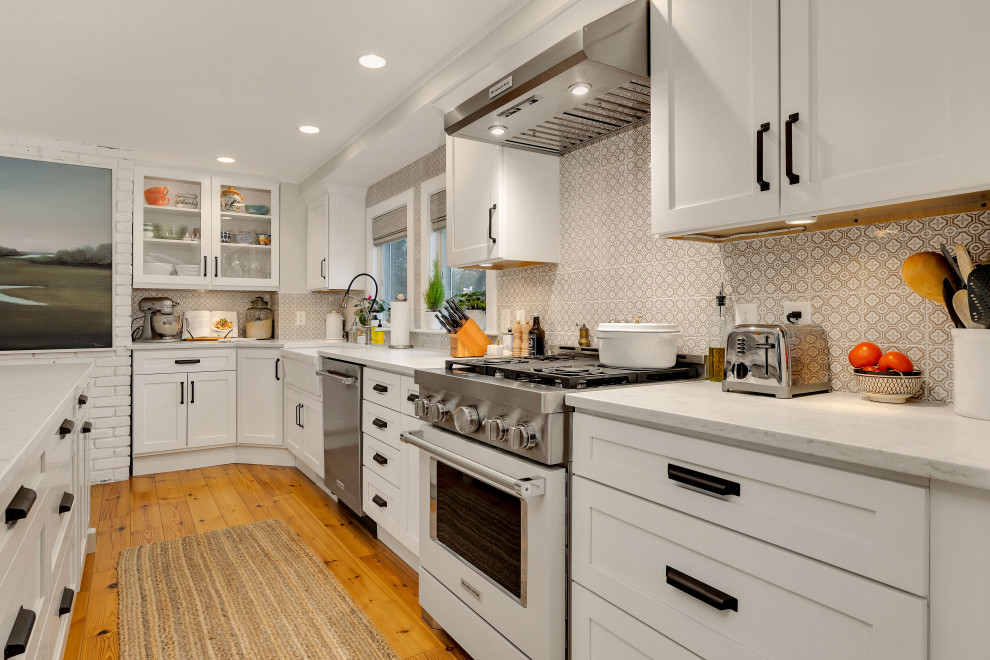 Inspiration for a large country l-shaped kitchen in Boston with a farmhouse sink, shaker cabinets, white cabinets, quartz benchtops, beige splashback, ceramic splashback, stainless steel appliances, light hardwood floors, with island, brown floor and white benchtop.