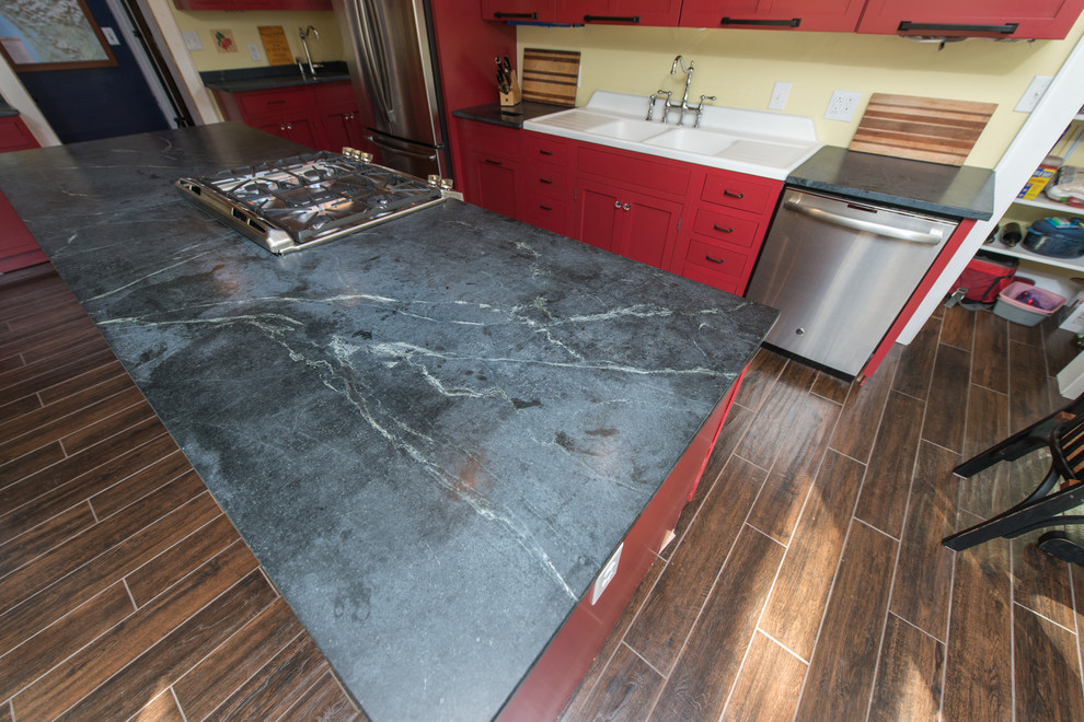 Mid-sized trendy galley dark wood floor and brown floor eat-in kitchen photo with a double-bowl sink, raised-panel cabinets, red cabinets, soapstone countertops, stainless steel appliances and an island