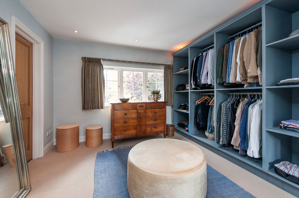 This is an example of a transitional men's dressing room in London with open cabinets, blue cabinets, carpet and beige floor.
