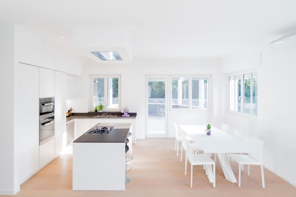 Large contemporary l-shaped open plan kitchen in Other with a single-bowl sink, flat-panel cabinets, white cabinets, white splashback, black appliances, light hardwood floors and with island.