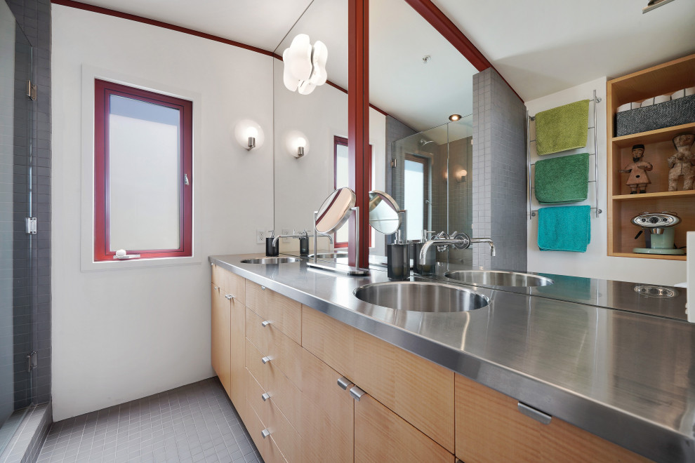 Photo of a mid-sized eclectic master bathroom in San Francisco with flat-panel cabinets, light wood cabinets, an alcove shower, a one-piece toilet, gray tile, ceramic tile, white walls, ceramic floors, an integrated sink, stainless steel benchtops, grey floor, a hinged shower door and grey benchtops.