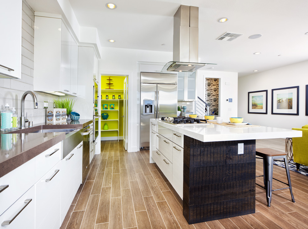 Contemporary l-shaped open plan kitchen in Orange County with a farmhouse sink, flat-panel cabinets, white cabinets, white splashback, subway tile splashback, stainless steel appliances and with island.