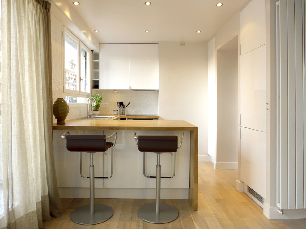Photo of a small contemporary u-shaped eat-in kitchen in Paris with a drop-in sink, flat-panel cabinets, white cabinets, wood benchtops, white splashback, light hardwood floors and a peninsula.