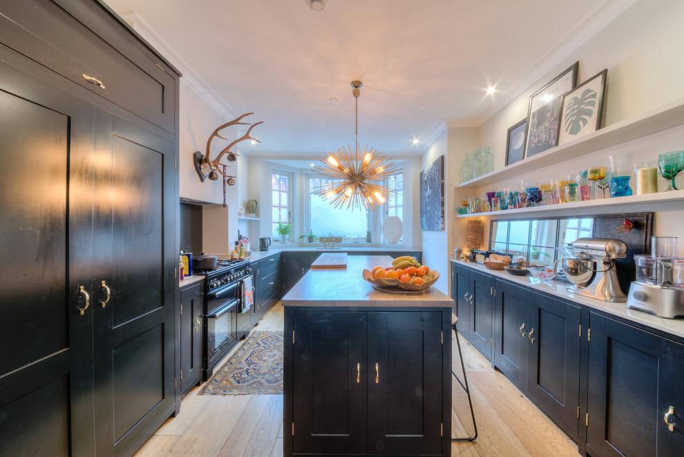 Photo of a large traditional galley open plan kitchen in London with an undermount sink, shaker cabinets, blue cabinets, wood benchtops, grey splashback, timber splashback, black appliances, light hardwood floors, with island, brown floor and white benchtop.