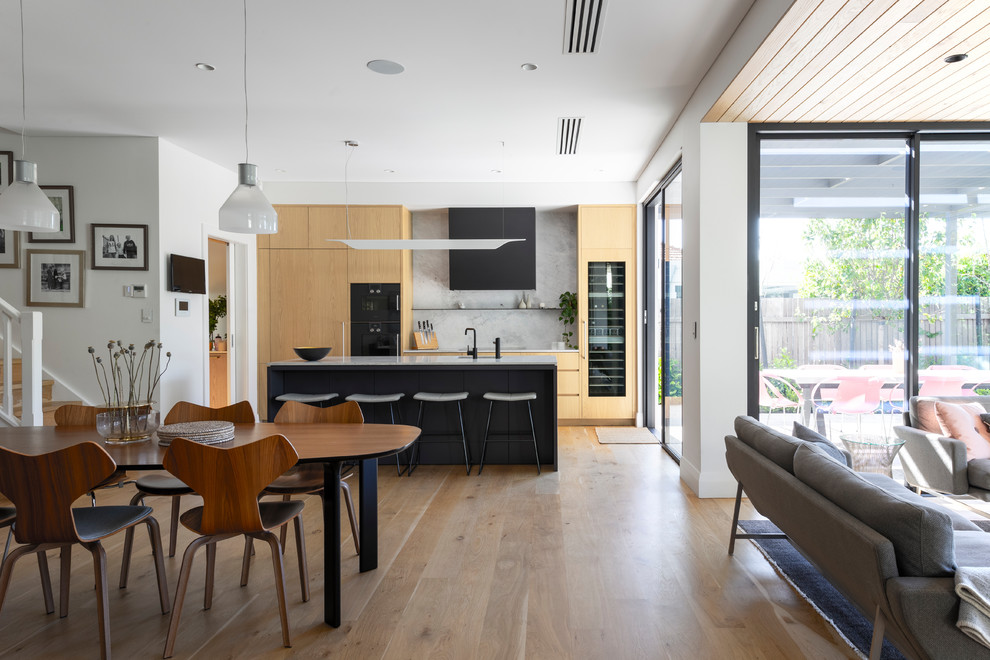 Contemporary galley open plan kitchen in Perth with a single-bowl sink, flat-panel cabinets, light wood cabinets, marble benchtops, grey splashback, marble splashback, black appliances, light hardwood floors, with island, grey benchtop and brown floor.