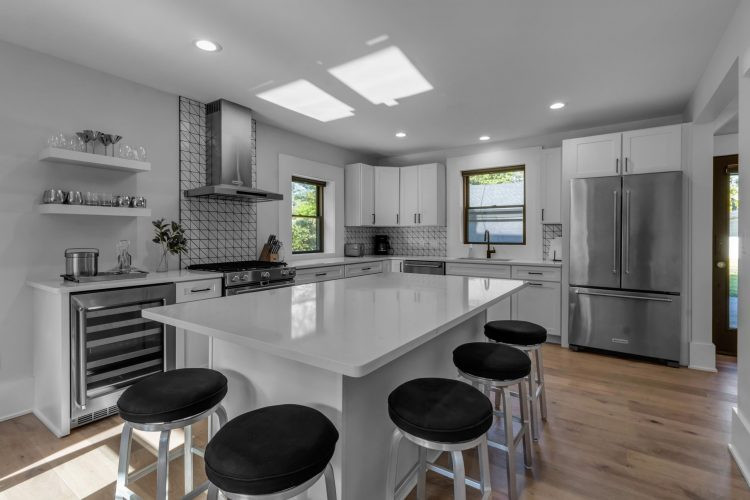 Mid-sized transitional l-shaped light wood floor and brown floor eat-in kitchen photo with an undermount sink, shaker cabinets, white cabinets, quartz countertops, white backsplash, ceramic backsplash, stainless steel appliances, an island and white countertops