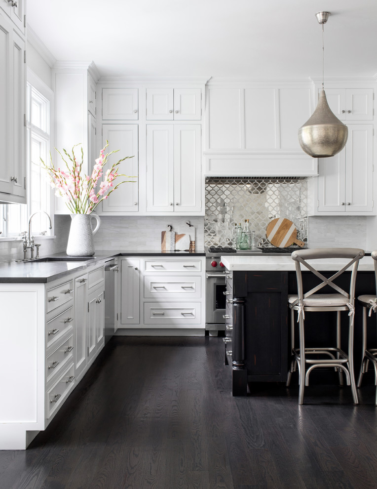 Beach style l-shaped kitchen in New York with white cabinets, metallic splashback, metal splashback, stainless steel appliances, dark hardwood floors, with island, black benchtop and beaded inset cabinets.