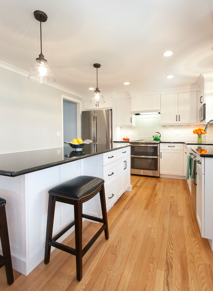 This is an example of a small traditional l-shaped eat-in kitchen in Bridgeport with a farmhouse sink, shaker cabinets, white cabinets, quartz benchtops, ceramic splashback, stainless steel appliances, light hardwood floors, with island, black benchtop and grey splashback.