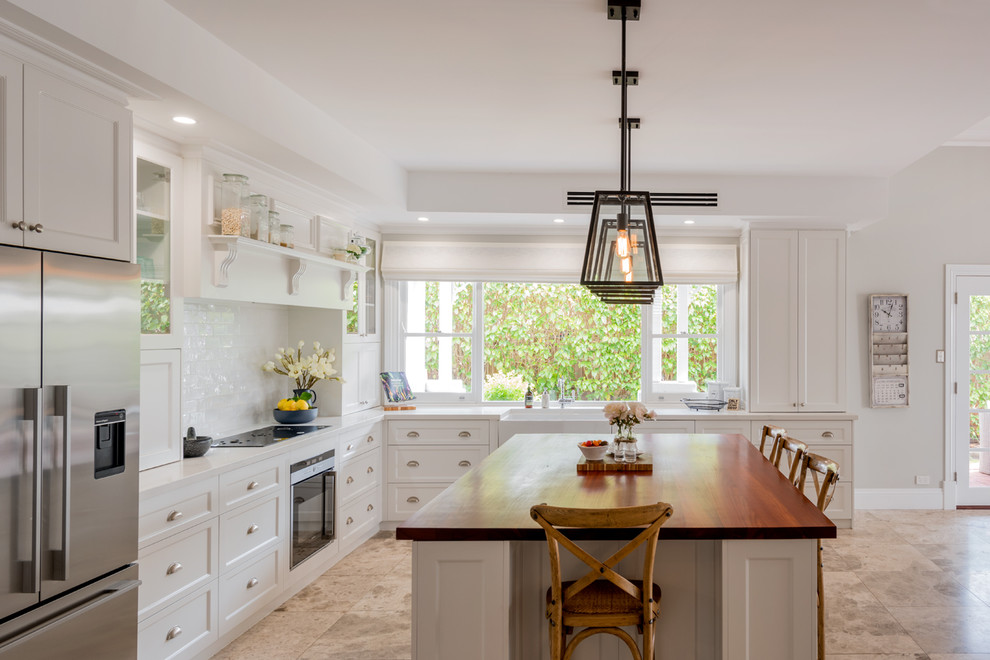 Design ideas for a large traditional l-shaped eat-in kitchen with a farmhouse sink, shaker cabinets, white cabinets, wood benchtops, white splashback, cement tile splashback, black appliances, porcelain floors, with island, beige floor and white benchtop.