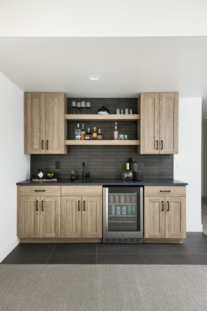 This is an example of a mid-sized transitional single-wall wet bar in Denver with an undermount sink, shaker cabinets, medium wood cabinets, quartz benchtops, black splashback, ceramic splashback, porcelain floors, black floor and black benchtop.
