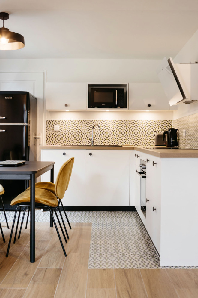 Foto på ett litet minimalistiskt brun kök, med en dubbel diskho, luckor med profilerade fronter, vita skåp, träbänkskiva, grönt stänkskydd, stänkskydd i keramik, svarta vitvaror, klinkergolv i keramik och beiget golv