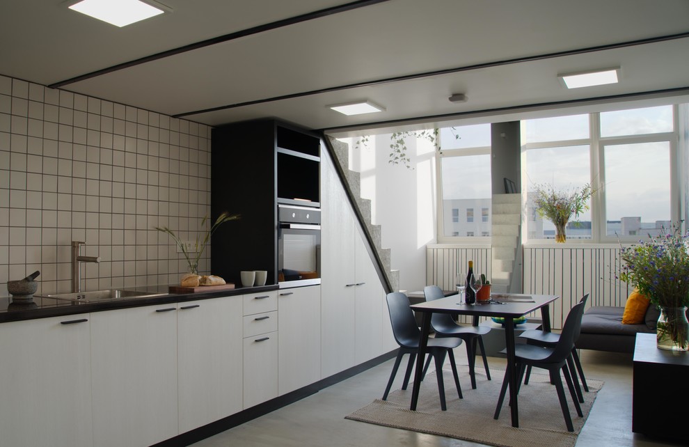 Design ideas for a mid-sized modern single-wall open plan kitchen in London with a drop-in sink, flat-panel cabinets, solid surface benchtops, white splashback, ceramic splashback, black appliances, concrete floors, no island, grey floor and black benchtop.