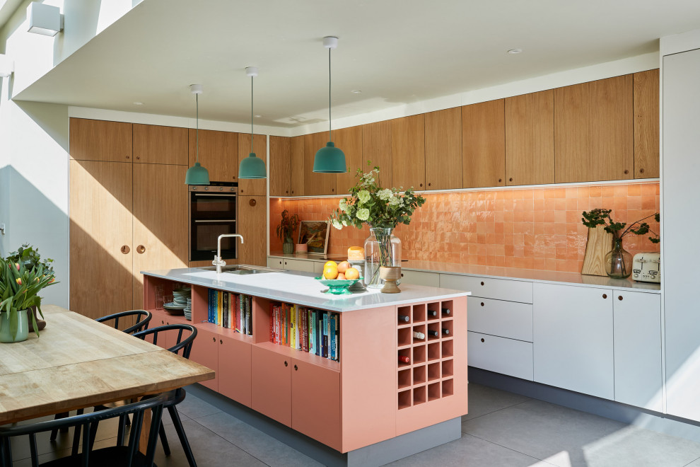 На фото: угловая кухня в стиле ретро с плоскими фасадами, фасадами цвета дерева среднего тона, оранжевым фартуком, островом, серым полом и белой столешницей