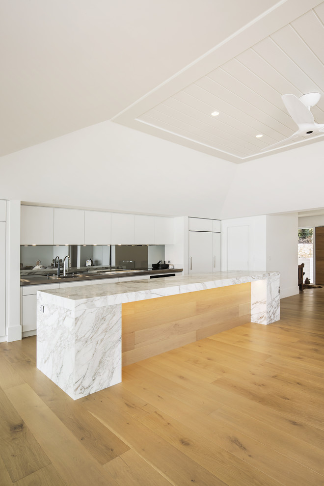 Inspiration for a large modern single-wall open plan kitchen in Sydney with an integrated sink, flat-panel cabinets, beige cabinets, marble benchtops, metallic splashback, mirror splashback, black appliances, light hardwood floors and with island.