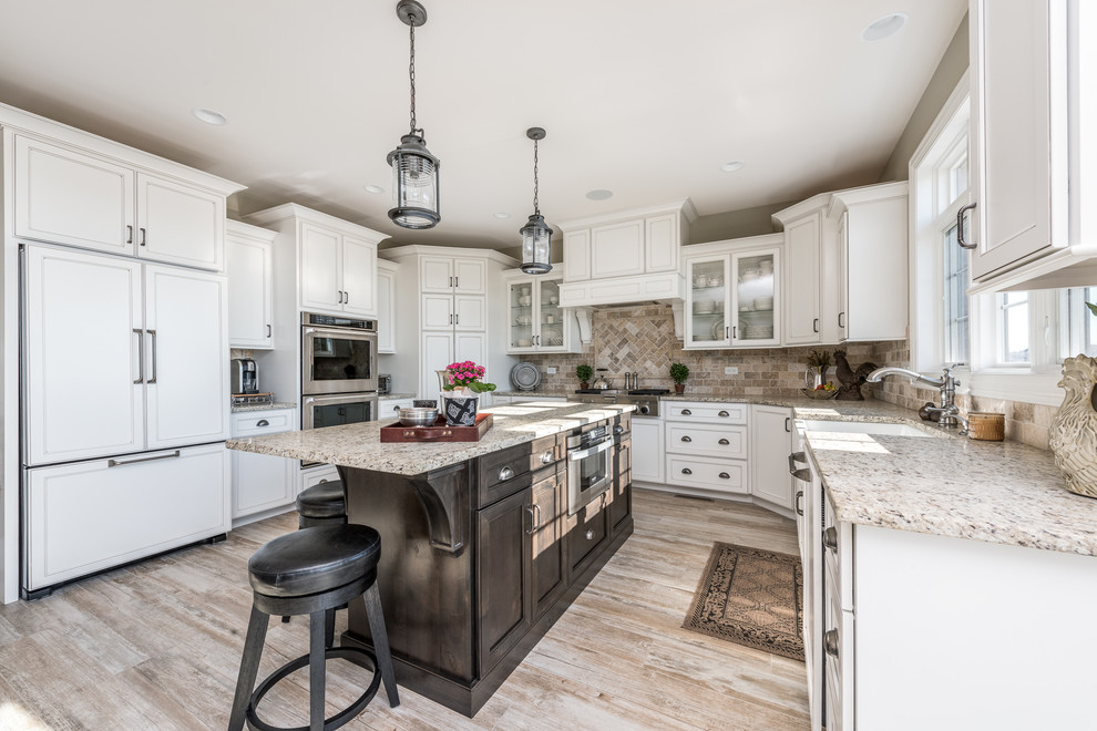Large traditional u-shaped open plan kitchen in Chicago with a farmhouse sink, recessed-panel cabinets, white cabinets, brown splashback, panelled appliances, light hardwood floors, with island, beige floor, beige benchtop, granite benchtops and stone tile splashback.