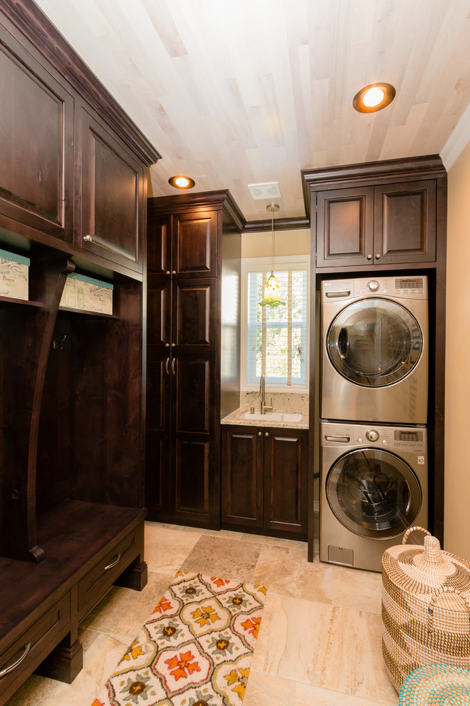 Photo of a small transitional l-shaped utility room in Minneapolis with an undermount sink, raised-panel cabinets, dark wood cabinets, granite benchtops, beige walls, travertine floors and a stacked washer and dryer.