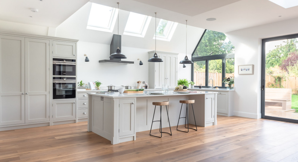 Large traditional u-shaped open plan kitchen in Surrey with shaker cabinets, white cabinets, quartzite benchtops, black appliances, light hardwood floors, with island, brown floor and white benchtop.