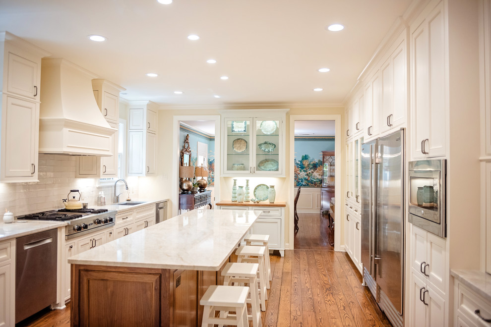 This is an example of a large transitional u-shaped eat-in kitchen in New Orleans with an undermount sink, white cabinets, white splashback, subway tile splashback, stainless steel appliances, with island, brown floor, white benchtop, medium hardwood floors, recessed-panel cabinets and quartzite benchtops.