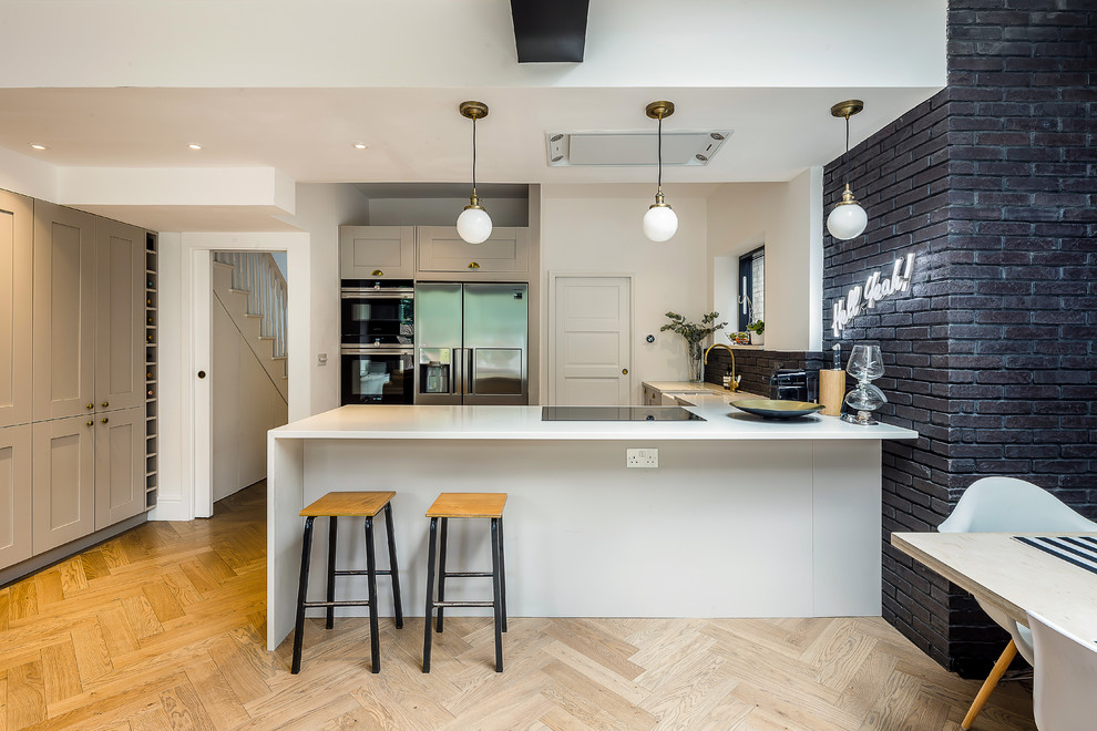 Example of a transitional u-shaped light wood floor and beige floor open concept kitchen design in London with a farmhouse sink, shaker cabinets, beige cabinets, black backsplash, brick backsplash, stainless steel appliances, a peninsula and white countertops