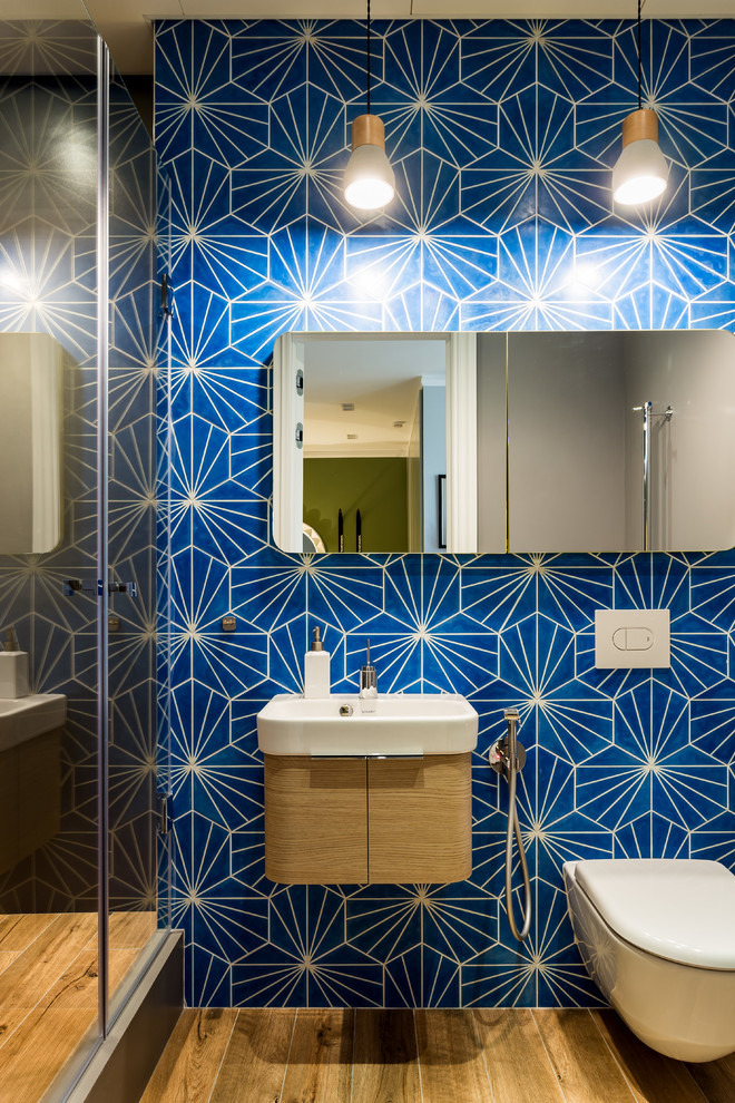Photo of a mid-sized contemporary 3/4 bathroom in Saint Petersburg with flat-panel cabinets, light wood cabinets, an alcove shower, a wall-mount toilet, blue tile, multi-coloured tile, white tile, ceramic tile, blue walls, ceramic floors, brown floor and a hinged shower door.