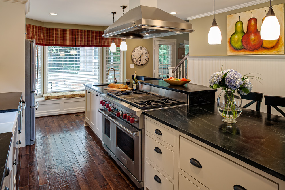 Photo of a small traditional galley eat-in kitchen in Charlotte with soapstone benchtops, a farmhouse sink, flat-panel cabinets, white cabinets, grey splashback, stone tile splashback, stainless steel appliances, dark hardwood floors and with island.