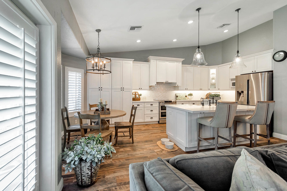 Large country l-shaped open plan kitchen in Phoenix with a farmhouse sink, shaker cabinets, white cabinets, quartz benchtops, white splashback, subway tile splashback, stainless steel appliances, with island and white benchtop.