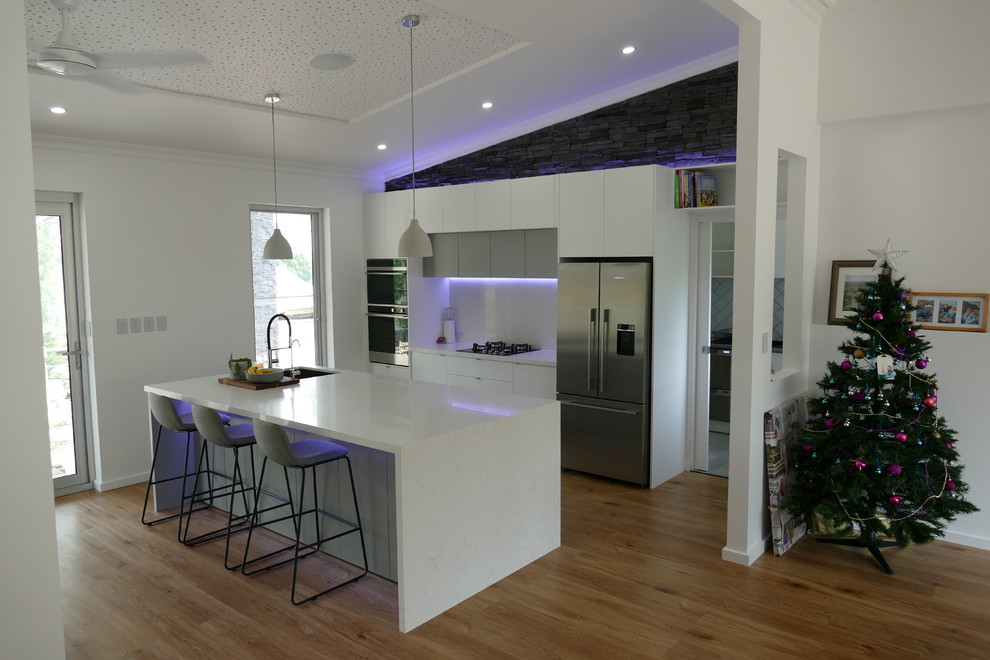 Photo of a mid-sized modern galley open plan kitchen in Adelaide with an undermount sink, flat-panel cabinets, white cabinets, quartz benchtops, white splashback, stone slab splashback, stainless steel appliances, medium hardwood floors, with island, brown floor and white benchtop.