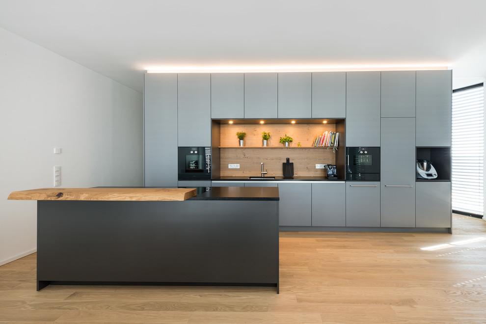 Large contemporary open plan kitchen in Munich with flat-panel cabinets, grey cabinets, brown splashback, timber splashback, black appliances, with island, brown floor, black benchtop and medium hardwood floors.