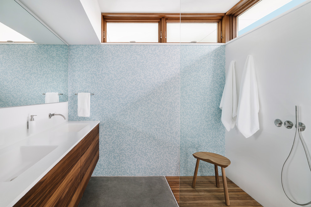 Photo of a contemporary master bathroom in Other with medium wood cabinets, an open shower, a wall-mount toilet, blue tile, porcelain tile, white walls, concrete floors, an integrated sink, solid surface benchtops, grey floor, an open shower and white benchtops.