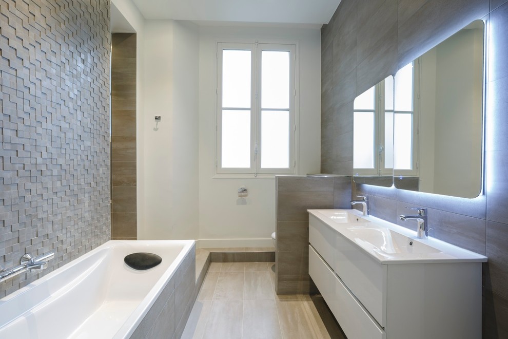 Mid-sized contemporary master bathroom in Paris with white cabinets, a drop-in tub, white walls, ceramic floors, an integrated sink, flat-panel cabinets, gray tile, a wall-mount toilet and stone tile.