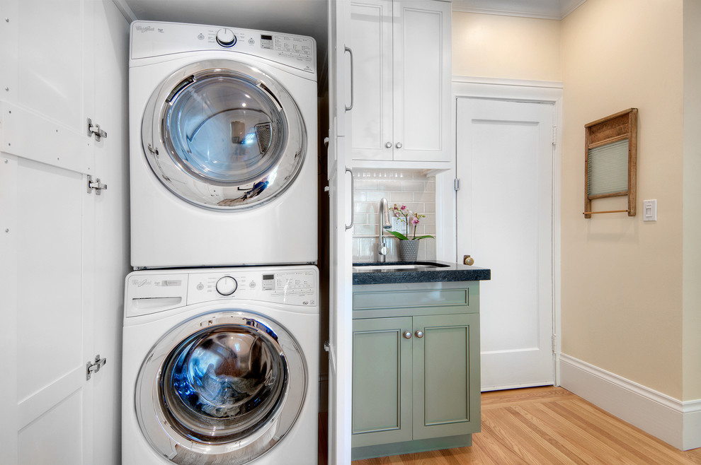 Design ideas for a small traditional single-wall laundry room in San Francisco with recessed-panel cabinets, white cabinets, granite benchtops, beige splashback, ceramic splashback, an undermount sink, beige walls, light hardwood floors and a stacked washer and dryer.