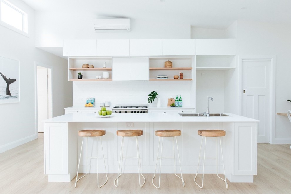 Photo of a beach style galley kitchen in Wollongong with a double-bowl sink, shaker cabinets, white cabinets, white splashback, stainless steel appliances, light hardwood floors, with island, beige floor and white benchtop.