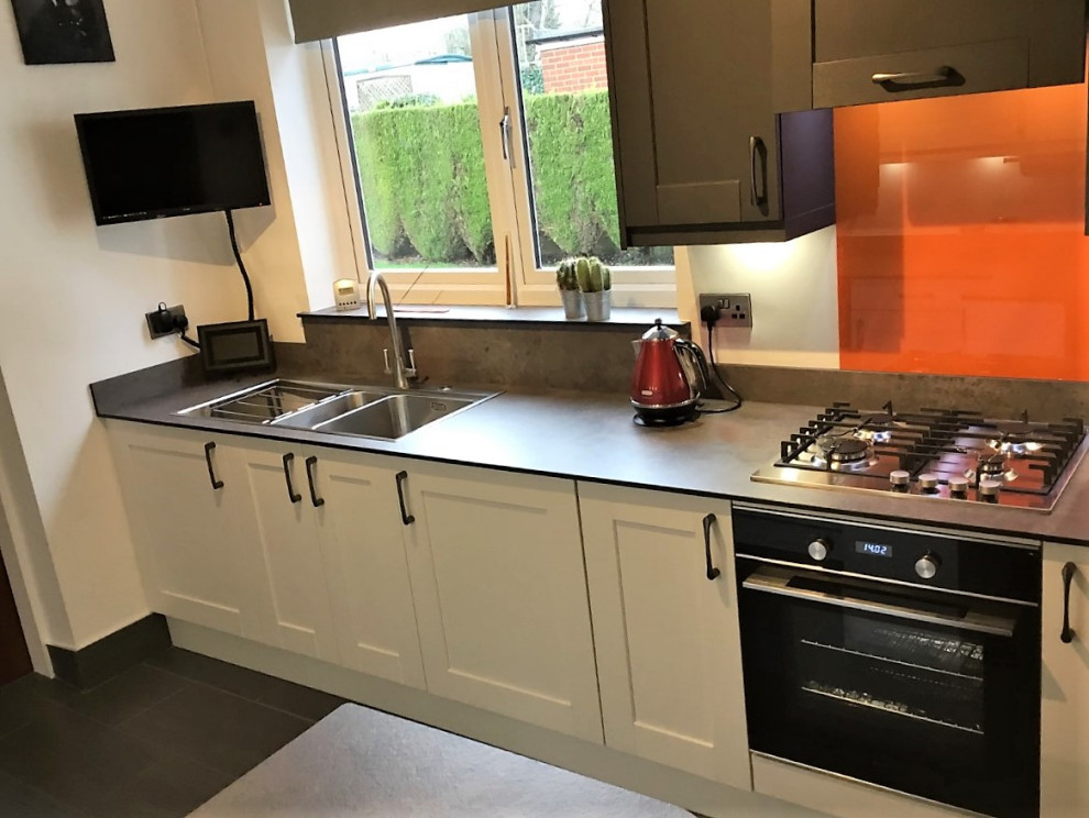 Cette photo montre une cuisine parallèle fermée et de taille moyenne avec un placard à porte shaker, une crédence orange, une crédence en feuille de verre, un sol en carrelage de porcelaine, une péninsule et un sol gris.