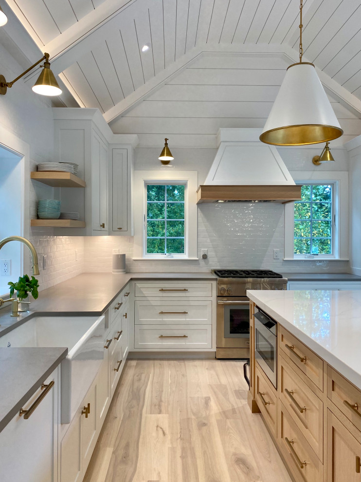 Beach style kitchen in Boston with a belfast sink, shaker cabinets, white cabinets, engineered stone countertops, white splashback, ceramic splashback, integrated appliances, medium hardwood flooring, an island, white worktops and a vaulted ceiling.