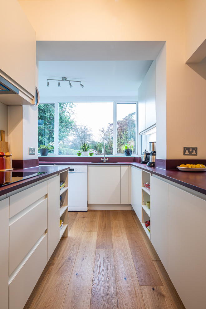 Design ideas for a small contemporary u-shaped kitchen/diner in Other with flat-panel cabinets, white cabinets, white splashback, glass sheet splashback, stainless steel appliances, no island, brown floors and red worktops.