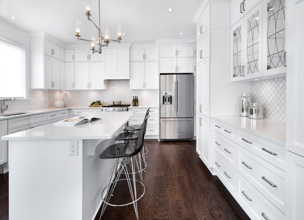 This is an example of a large transitional u-shaped open plan kitchen in Ottawa with an undermount sink, shaker cabinets, white cabinets, quartzite benchtops, white splashback, ceramic splashback, white appliances, dark hardwood floors and with island.