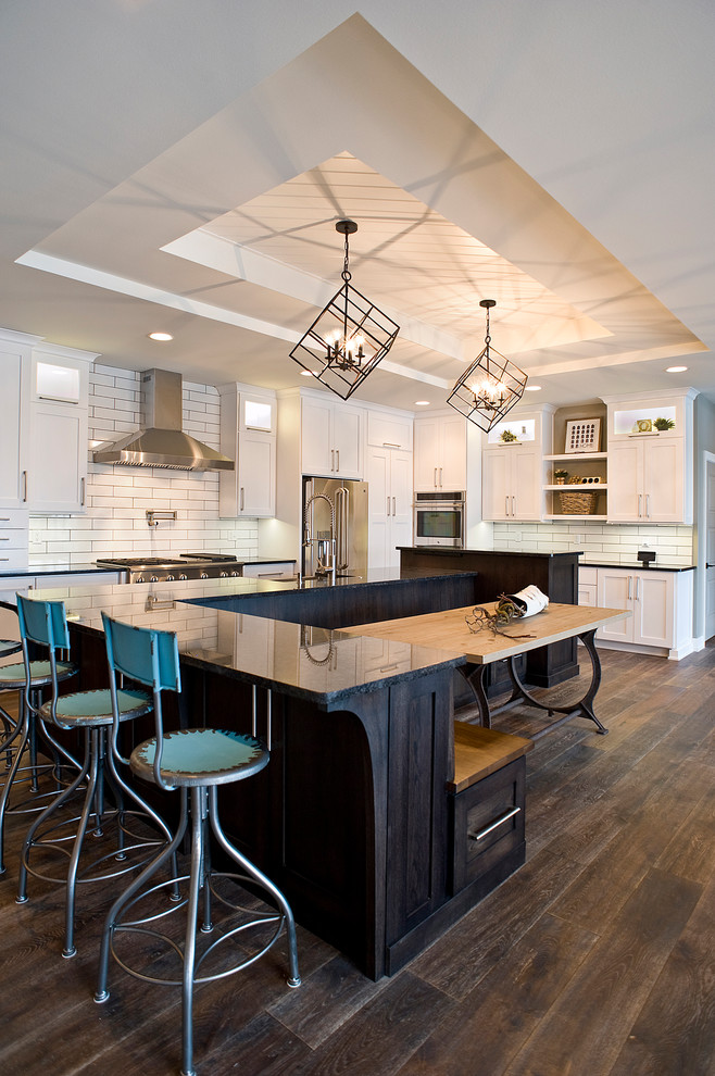 Large transitional l-shaped dark wood floor and brown floor eat-in kitchen photo with a farmhouse sink, raised-panel cabinets, white cabinets, quartz countertops, white backsplash, subway tile backsplash, stainless steel appliances and an island