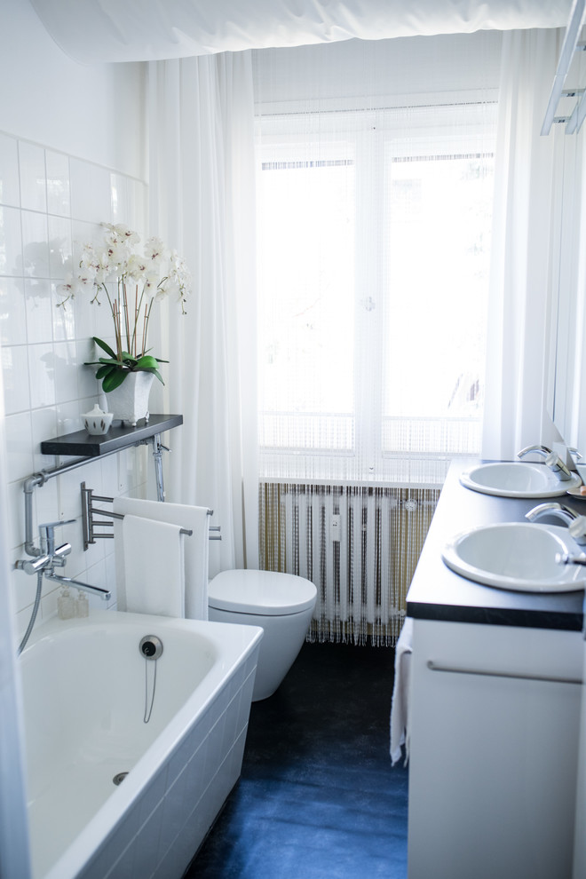 Small contemporary 3/4 bathroom in Berlin with white cabinets, ceramic tile, white walls, an integrated sink, black floor, black benchtops, a drop-in tub, a shower/bathtub combo, a wall-mount toilet and white tile.