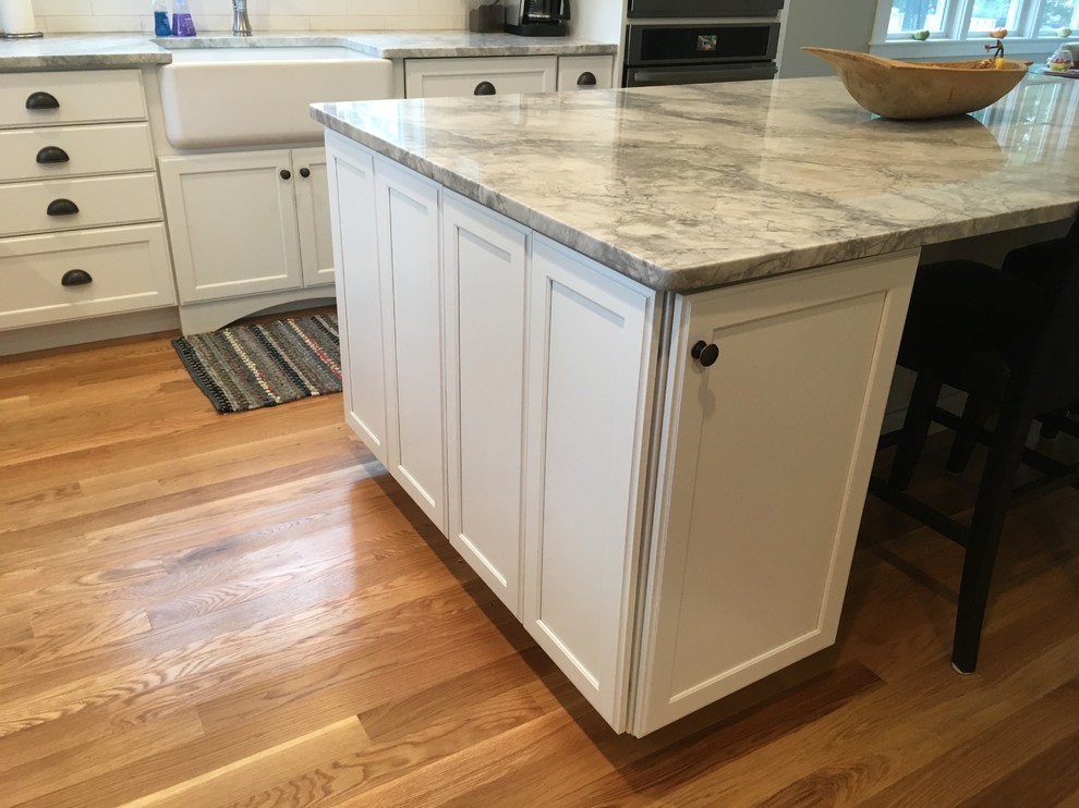 Inspiration for a transitional l-shaped medium tone wood floor eat-in kitchen remodel in Boston with a farmhouse sink, shaker cabinets, white cabinets, white backsplash, porcelain backsplash, stainless steel appliances and an island