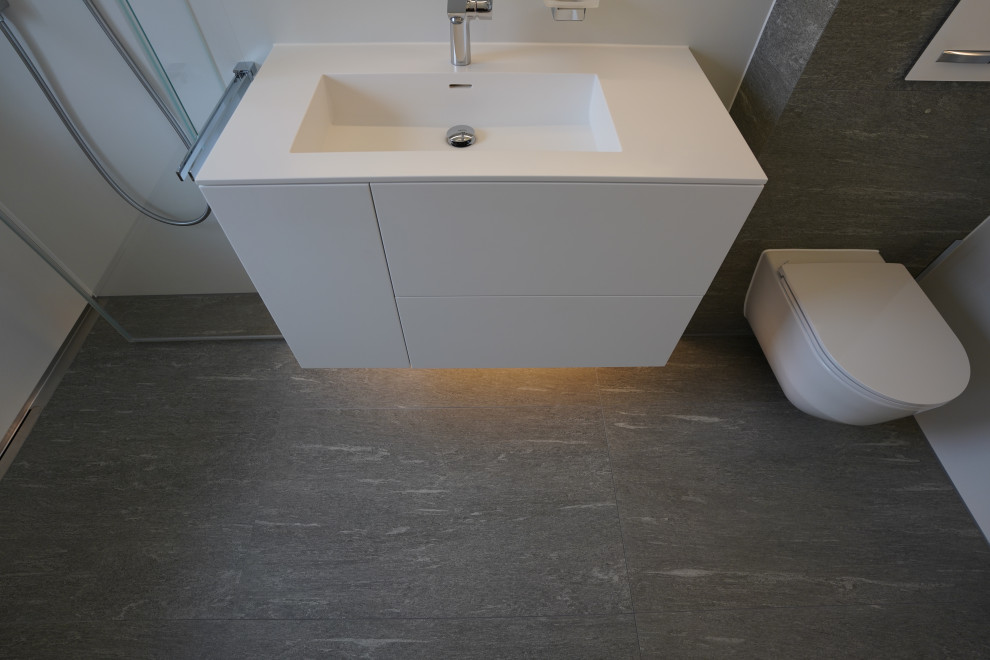 Duschbad With flächenbündigen Schrankfronten, weißen Schränken, bodengleicher Dusche, Wandtoilette, weißen Fliesen, Steinfliesen, Wandwaschbecken, Mineralwerkstoff-Waschtisch, grauem Boden, Falttür-Duschabtrennung, weißer Waschtischplatte, Einzelwaschbecken and schwebendem Waschtisch in Munich