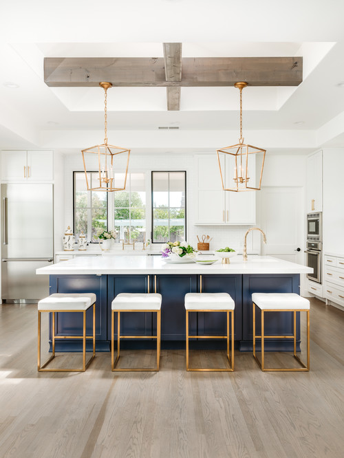 Bright white kitchen with a navy blue island kitchen cabinet, gold pendant lights and four upholstered stools on light wood flooring.
